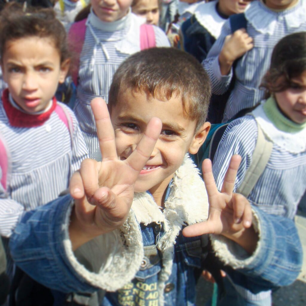 Smiling Gazan schoolchild holding up hands showing five fingers in a crowd of other children